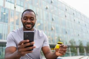 afrikansk amerikan man handla uppkopplad innehav smartphone betalande med guld kreditera kort kille på urban gata i stad uppköp på internet stiga på kreditera kort detaljer uppkopplad handla e-handel leverans service foto