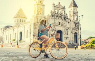 Lycklig flicka i hatt ridning en cykel och textning med de telefon. Lycklig turist flicka i hatt på cykel använder sig av telefon på de gata. granada, nicaragua foto