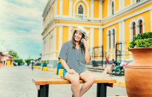 leende turist Sammanträde på en bänk talande på cell telefon. Lycklig resa kvinna Sammanträde kallelse på de telefon i en turist fyrkant foto