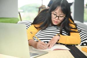 tonårs kvinnlig student sover på kuddar läser bok och använder bärbar dator. hon skriver anteckningar för att förbereda sig för tentamen.