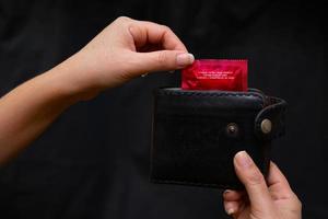närbild kvinna hand håller svart läder plånbok med en röd kondom på den svarta bakgrunden foto