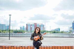 spelar ukulele av ung skön asiatisk kvinna bär jacka och svart jeans Framställ utomhus foto