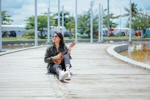 spelar ukulele av ung skön asiatisk kvinna bär jacka och svart jeans Framställ utomhus foto