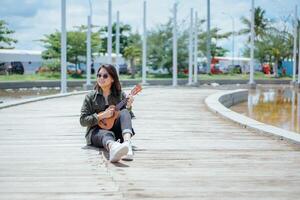 spelar ukulele av ung skön asiatisk kvinna bär jacka och svart jeans Framställ utomhus foto