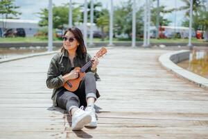 spelar ukulele av ung skön asiatisk kvinna bär jacka och svart jeans Framställ utomhus foto