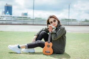 spelar ukulele av ung skön asiatisk kvinna bär jacka och svart jeans Framställ utomhus foto