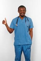 leende svart kirurg läkare skäggig man i blå täcka med stetoskop gör tummen upp tycka om gest foto