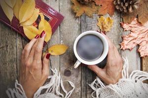 varmt kaffe i händerna på en tjej, en bok med dikter, höstlöv, en stickad tröja på en träbordsbakgrund. mysig höststämning i oktober, november foto