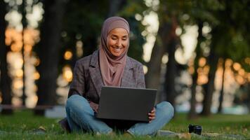 Lycklig leende muslim islamic företag kvinna flicka kvinna studerande bär hijab Sammanträde på grön gräs gräsmatta i stad parkera använder sig av bärbar dator för avlägsen e-learning arbete chattar uppkopplad kontroll post med kaffe foto