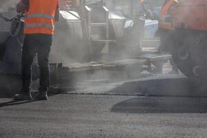 män arbetssätt med asfalte utläggare maskin under väg gata reparation Arbetar på dag ljus med rök och ånga i de luft foto
