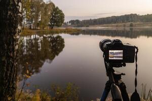 svart digital kamera på stativ skytte tidigt dimmig morgon- landskap på höst sjö med selektiv fokus foto