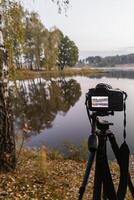 svart digital kamera på stativ skytte tidigt dimmig morgon- landskap på höst sjö med selektiv fokus foto