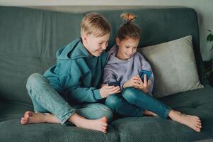 två barn Sammanträde på en soffa spelar med deras telefoner foto