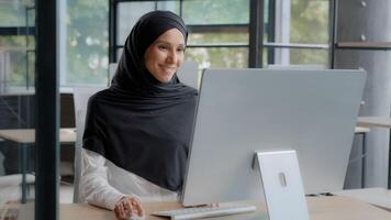 Lycklig framgångsrik arab affärskvinna i hijab skriver på tangentbord arbetssätt på dator njuter arbete uppkopplad i modern kontor leende kvinna chef placering beställa på internet kommunicerar på social media foto