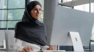 ung arab affärskvinna i hijab arbetssätt på dator leende njuter kontor arbete framgångsrik kvinna professionell chef företag anställd utvecklas projekt uppkopplad sökande för information på internet foto