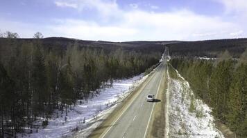 flygande över de betong väg med rör på sig fordon längs de tall träd skog. klämma. antenn se av snö täckt jord i de skog och sent höst Land väg med en körning bil. foto