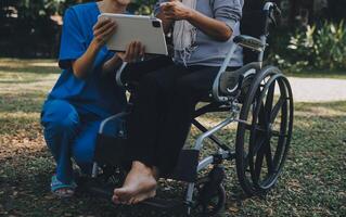 äldre asiatisk senior kvinna på rullstol med asiatisk försiktig vårdgivare och uppmuntra patient, gående i trädgård. med vård från en vårdgivare och senior hälsa försäkring i Sol ljus foto