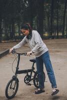 Lycklig asiatisk ung kvinna promenad och rida cykel i parkera, gata stad henne leende använder sig av cykel av transport, eco vänlig, människor livsstil begrepp. foto