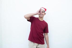 ung asiatisk man bär en santa claus hatt med en hand framställning de ok gest isolerat förbi en vit bakgrund för visuell kommunikation foto