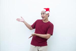 ung asiatisk man bär en santa claus hatt leende, chock och pekande till henne sida isolerat förbi vit bakgrund för visuell kommunikation foto