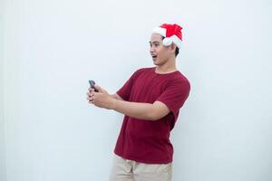 ung asiatisk man bär en santa claus hatt innehav en smartphone och uttrycker en leende, chock och överraskning isolerat förbi en vit bakgrund för visuell kommunikation foto