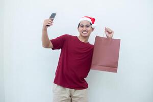 ung asiatisk man bär en santa claus hatt innehav en smartphone och en handla väska med uttryck av leende, chock, och överraskning, isolerat mot en vit bakgrund för visuell kommunikation foto
