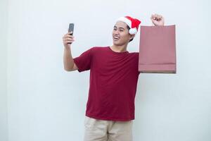 ung asiatisk man bär en santa claus hatt innehav en smartphone och en handla väska med uttryck av leende, chock, och överraskning, isolerat mot en vit bakgrund för visuell kommunikation foto