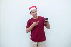 ung asiatisk man bär en santa claus hatt innehav en smartphone och uttrycker en leende, överraskning, och pekande till de sida, isolerat förbi en vit bakgrund för visuell kommunikation foto