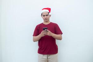 ung asiatisk man bär en santa claus hatt innehav en smartphone och uttrycker en leende, chock och överraskning isolerat förbi en vit bakgrund för visuell kommunikation foto