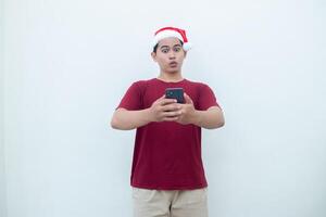 ung asiatisk man bär en santa claus hatt innehav en smartphone och uttrycker en leende, chock och överraskning isolerat förbi en vit bakgrund för visuell kommunikation foto