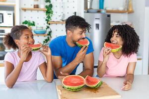 ung familj äter vattenmelon och har roligt. blandad lopp familj i kök tillsammans äter en vattenmelon skiva. foto