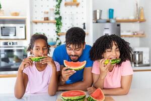 ung familj äter vattenmelon och har roligt. blandad lopp familj i kök tillsammans äter en vattenmelon skiva. foto