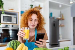 leende Söt kvinna ser på mobil telefon och innehav glas av orange juice medan matlagning färsk grönsaker i kök interiör på Hem foto