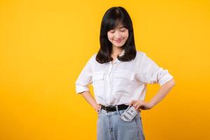 fängslande bild av asiatisk ung kvinna bär vit skjorta och denim jeans, självsäkert innehav kontanter från henne skjorta ficka. mode möter finansiell bemyndigande. perfekt för finansiera och mode teman. foto