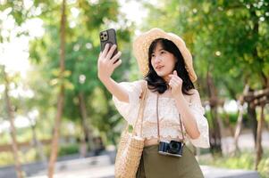 porträtt av asiatisk ung kvinna resande med vävning hatt, korg, mobil telefon och kamera på grön offentlig parkera bakgrund. resa resa livsstil, värld resa explorer eller Asien sommar turism begrepp. foto
