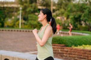 kvinna joggare. passa ung asiatisk kvinna med grön sportkläder aerobics dansa övning i parkera och njuter en friska utomhus. kondition löpare flicka i offentlig parkera. wellness varelse begrepp foto