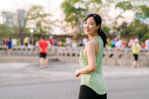 kvinna joggare. passa ung asiatisk kvinna med grön sportkläder aerobics dansa övning i parkera och njuter en friska utomhus. kondition löpare flicka i offentlig parkera. wellness varelse begrepp foto