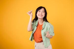känna de kärlek i de luft med härlig ung asiatisk kvinna i henne 30-talet. hon älskvärt visas en mini hjärta gest och en mild leende. kropp språk begrepp. bär ett orange skjorta och grön hoppare. foto