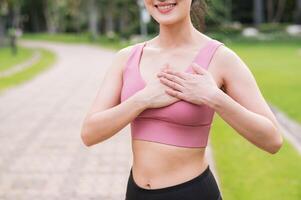 kvinna joggare. ung asiatisk kvinna Lycklig leende bär rosa sportkläder innehav händer på bröst innan löpning i offentlig parkera. sjukvård wellness begrepp. foto