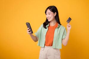 erfarenhet de spänning av mobil handla med en vibrerande ung asiatisk kvinna i henne 30-talet, påklädning orange skjorta och grön hoppare, använder sig av smartphone medan presenter kreditera kort på gul studio bakgrund. foto