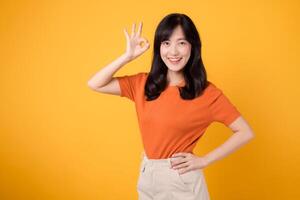 självsäker ung asiatisk kvinna i henne 30s bär orange skjorta, visning Okej tecken på vibrerande gul bakgrund. positiv hand gest begrepp. foto