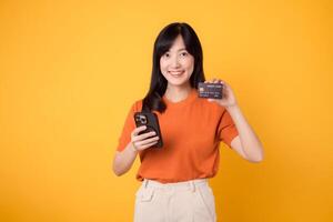 entusiastisk ung asiatisk kvinna i henne 30-talet, använder sig av smartphone och innehav kreditera kort på gul bakgrund. snabb uppkopplad handla. foto