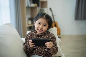 asiatisk barn flicka ser använder sig av och Rör mobil telefon skärm på soffa soffa. bebis leende rolig tid till använda sig av mobil telefon. för mycket skärm tid. söt flicka tittar på videoklipp medan tv, internet missbruk. foto