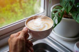 personens hand är innehav ett aromatisk latte konst kaffe kopp. dryck kaffe på de morgon- nära de fönster foto