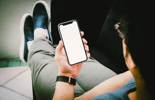 man innehav en smartphone med vit skärm mock-up, koppla av Sammanträde på en soffa i de levande rum på Hem foto