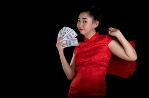 porträtt av en vacker ung asiatisk kvinna röd klänning traditionell cheongsam håller en väska shopping och pengar sedel 100 usd på den svarta bakgrunden foto