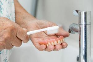 asiatisk senior eller äldre gammal kvinna patienten använder tandborste för att rengöra delvis protes av ersättningständer.