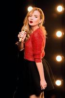 ung smiley vackert långt hår i röd tröja flicka med mikrofon sjunger sång på scenen i karaoke foto