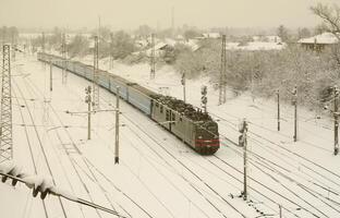 en lång tåg av passagerare bilar är rör på sig längs de järnväg Spår. järnväg landskap i vinter- efter snöfall foto