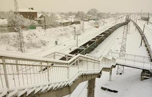 en lång tåg av frakt bilar är rör på sig längs de järnväg Spår. järnväg landskap i vinter- efter snöfall foto